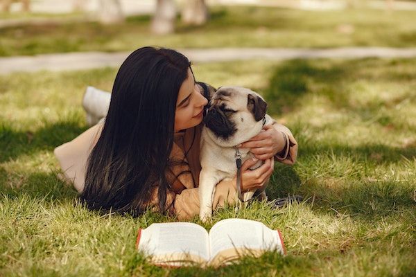 pug being hugged by a woman with long brown hair in the grass with an open book in front of them