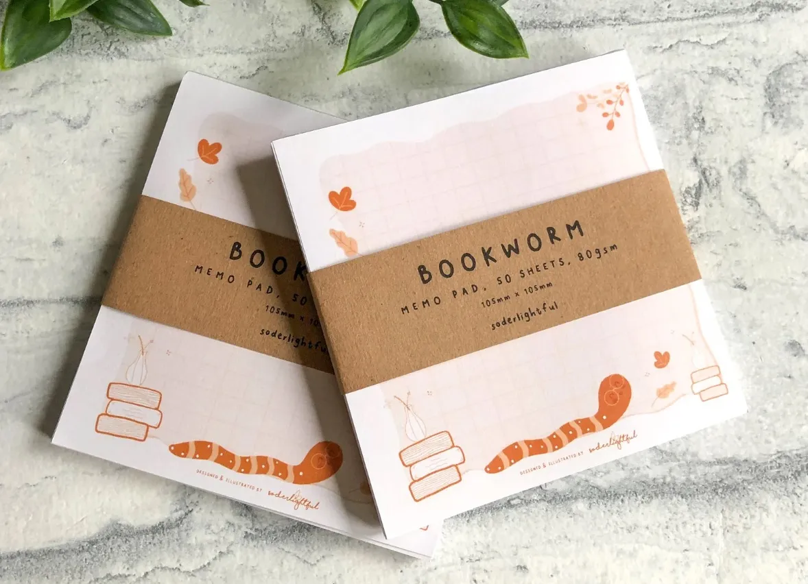 Image of two notepads featuring an orange and creme color palette. They have stacks of books and little worms. 