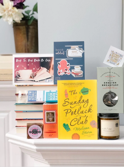 a photo of a Sweets Reads Box, showing The Sunday Potluck club surrounded by bookmarks, tea, a candle, and more goodies