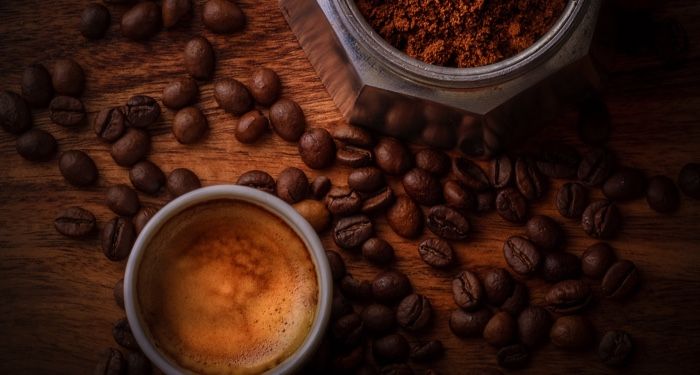 coffee beans scattered on a wood surface between a white ceramic mug full of coffee and the bottom half of a moka pot filled with ground coffee