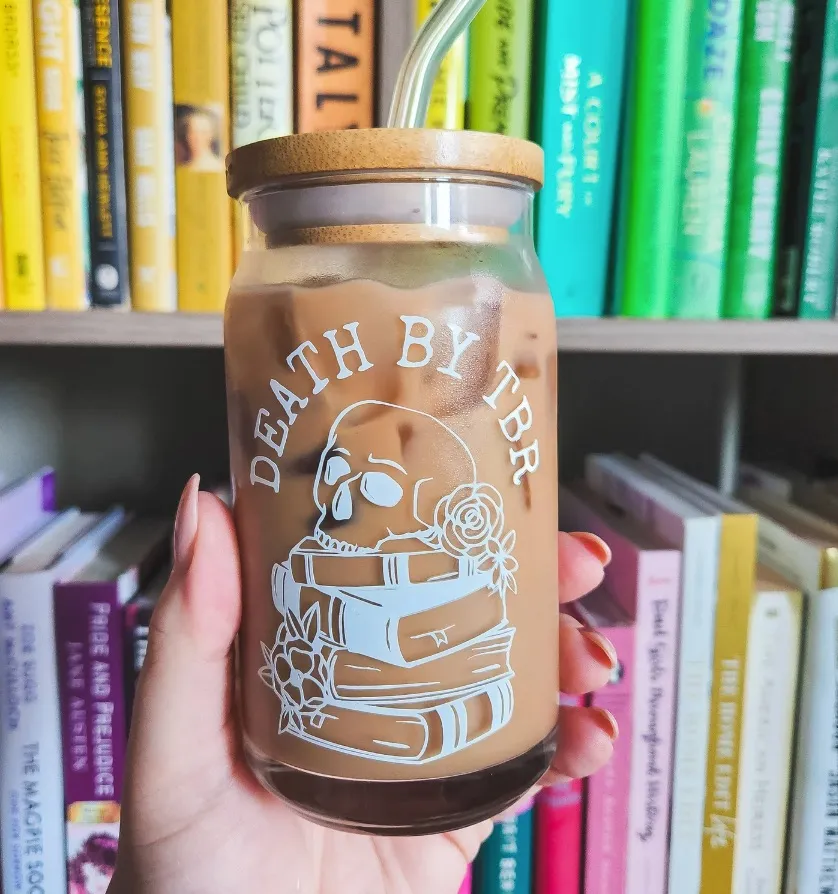 Image of a glass with a lid held by a white hand in front of bookshelves. In white on the glass are the words "death by tbr," with an image of a skull and books.