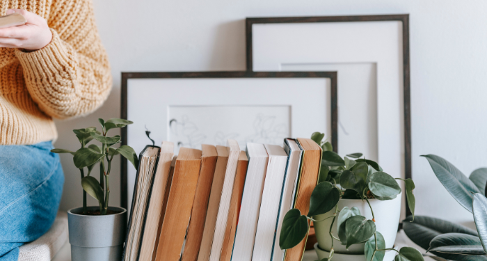 a photo of a bookshelf with plants. The books are shelved backwards. To the side is a person mostly cropped out of the photo who is reading