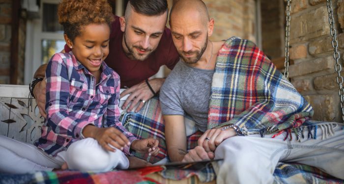 queer family with two fair-skinned dads and a brown-skinned child reading a book and smiling