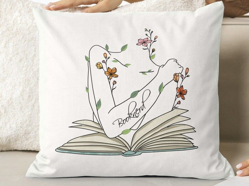 Photo of a white pillow with a print of an open book and flowers coming out of it. the text in the centre says booknerd.