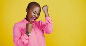 person with brown skin cheering with both hands held in the air. They are wearing a pink button-down shirt and standing against a bright yellow background