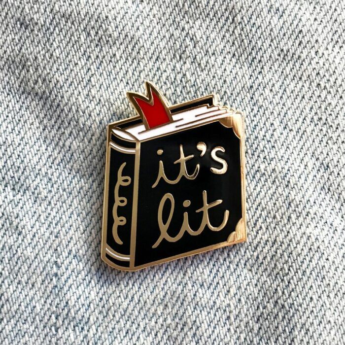 enamel pin in the shape of a black book with gold cursive words saying "it's lit"