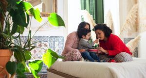 two women with brown skin sitting on a couch feeding a toddler