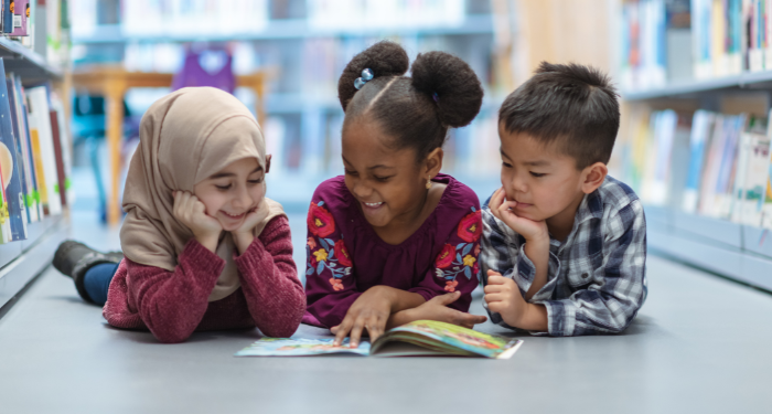 a photo of three kids on the floor reading a book together and smiling