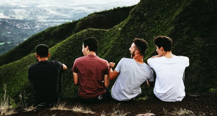 four men with lighter skin tones sitting on the ground, and looking over lush, green hills and laughing