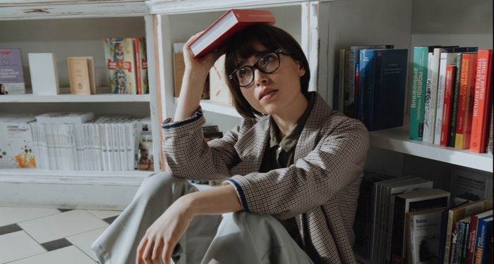 fair-skinned woman sitting in front of bookshelf, holding a book on her head