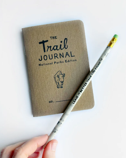 A small brown journal that says "Trail Journal: National Parks" in a vintage font, along with a hand holding a pencil that says "Leave it better than you found it"