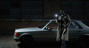 brown-skinned Black man in trench coat standing by a car holding a flashlight
