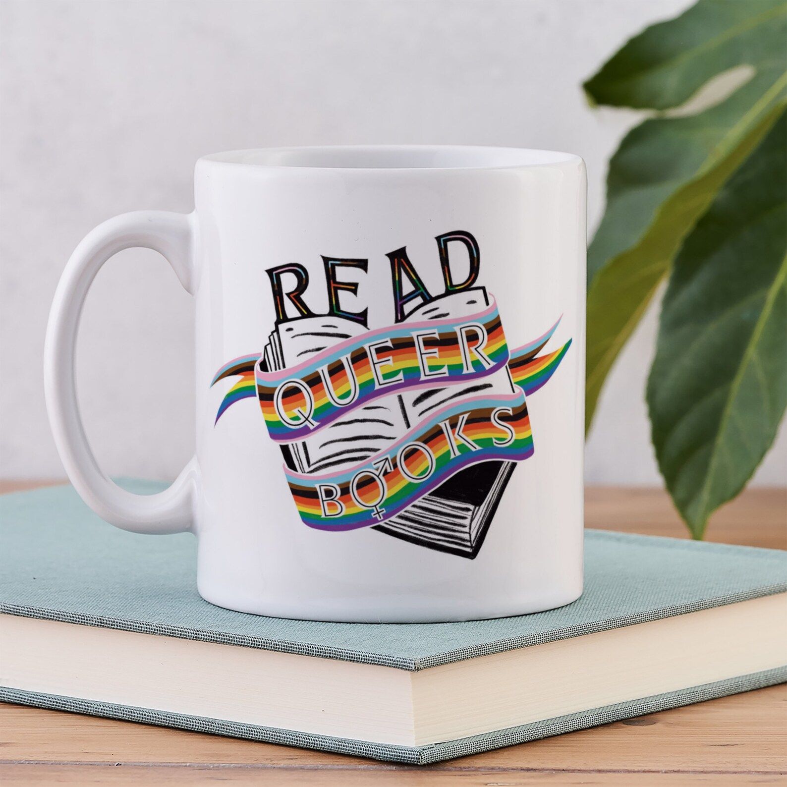 Image of a white mug with the words "read queer books" with an open book and rainbow flag. 