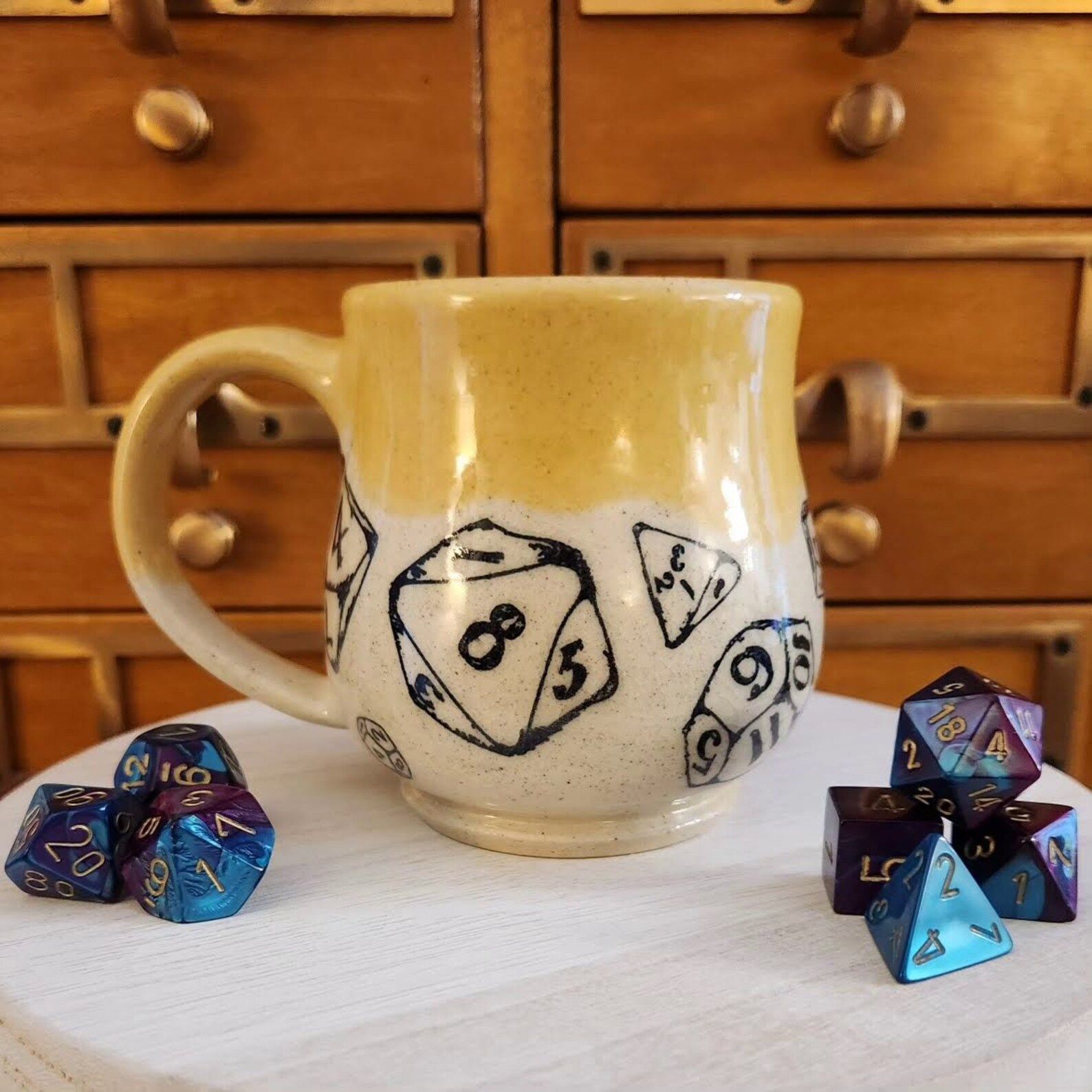 A Yellow and white handmade Ceramic Mug with dice painted on the bottom half sits on a table with a set of 7 dnd dice in front of woden drawers. 