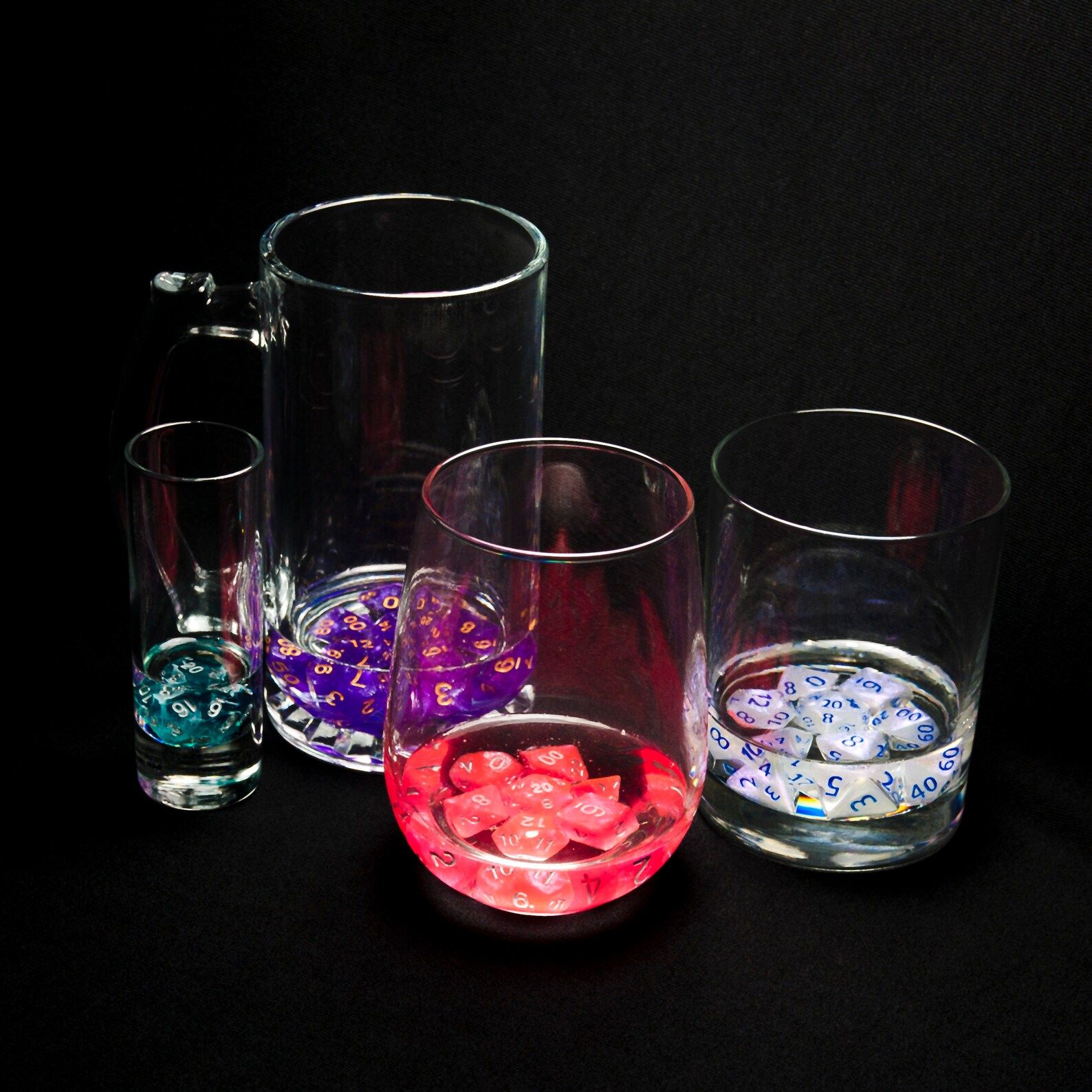 Four glasses on a black background. A shot glass with a green die, a mug with purple dice, a stemless wine glass with red dice and a whisky glass with white dice from left to right.,