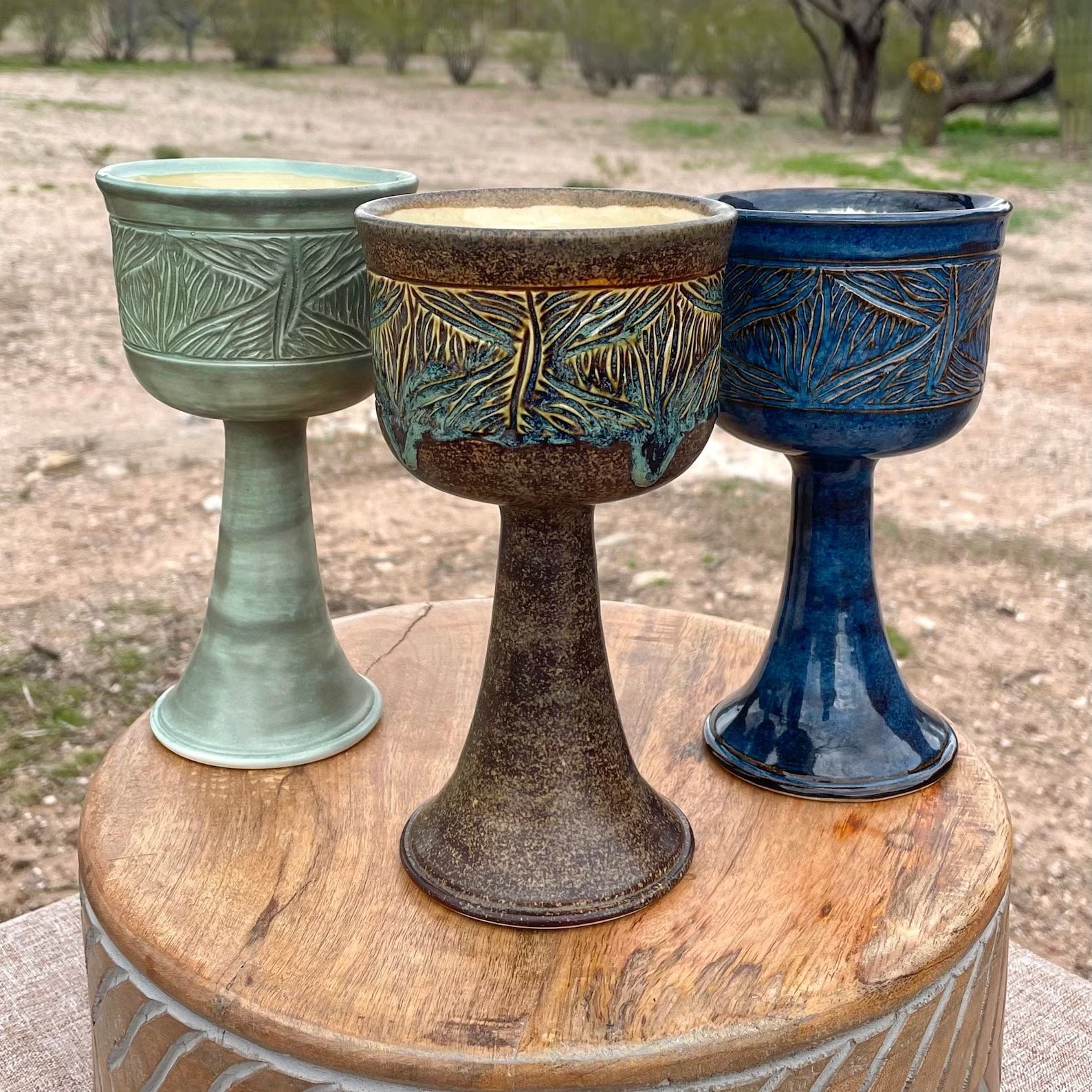 A green, brown, and blue ceramic goblet sit on a wooden table in front of a yard. 