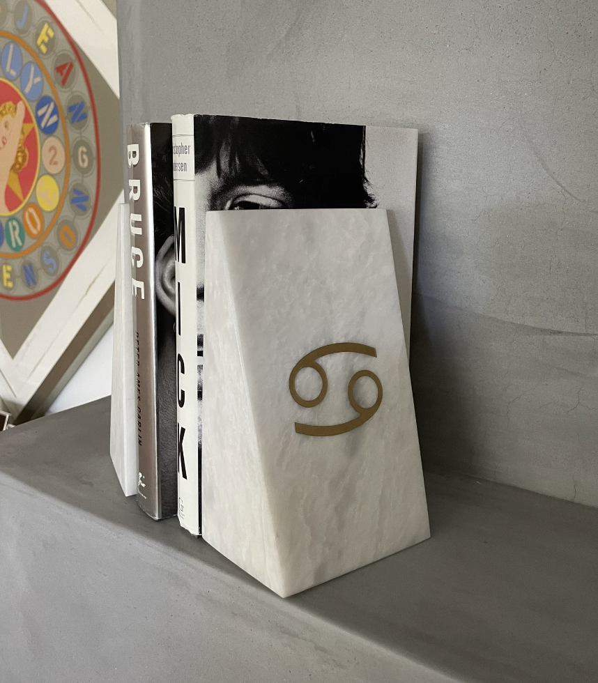 A white marble triangular bookend with an engraved Cancer symbol holding up a few books on a gray mantle