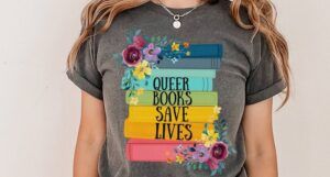 A photo of someone wearing a tee shirt with an illustration of a stack of rainbow colored book spines, florals, and the words "Queer books save lives."