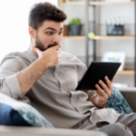 a photo of a man reading on a tablet with eyebrows raised and a hand over his mouth