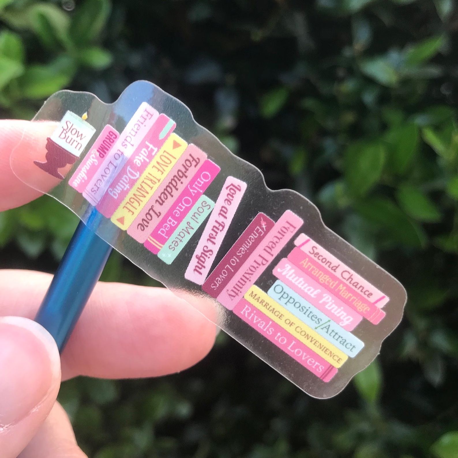 A Romance Tropes Pink Bookshelf sticker with a clear background in front of a bush, a hand, and a pen.