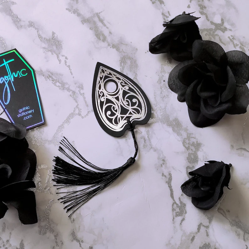 Photo of a bookmark resembling an Ouija Board planchette placed on a surface with black roses next to i. 