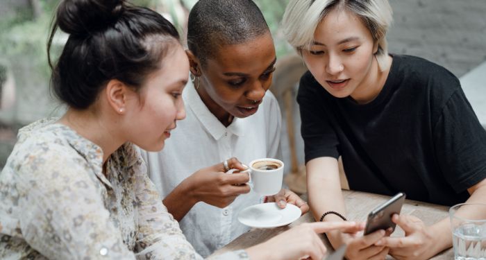 two fair-skinned Asian women and one brown0skinned Black woman looking at a cell phone with great interest