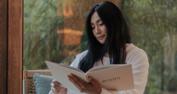 beige-skinned Asian woman sitting and reading a book with pne trees in the background