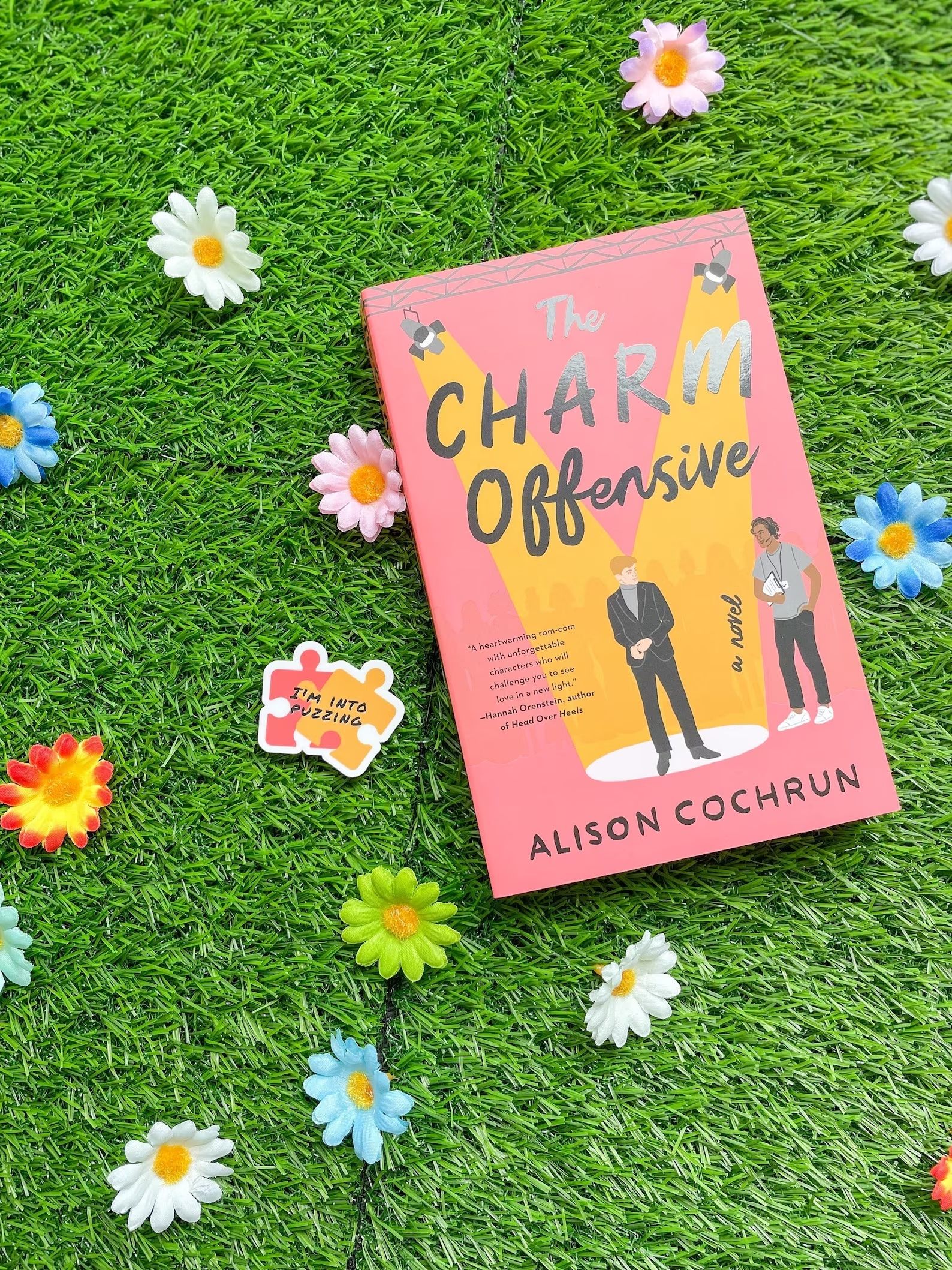 On top of grass and flowers is a sticker made to look like the cover of the book the charm offensive by Alison Cochrun 