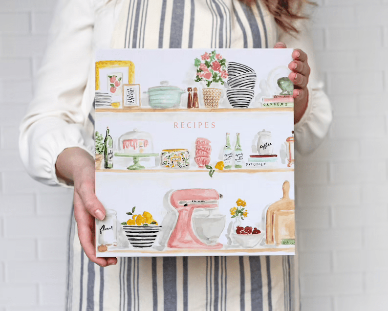 Photo of a woman holding a recipe notebook binder with a cover of painted shelves with kitchen tools