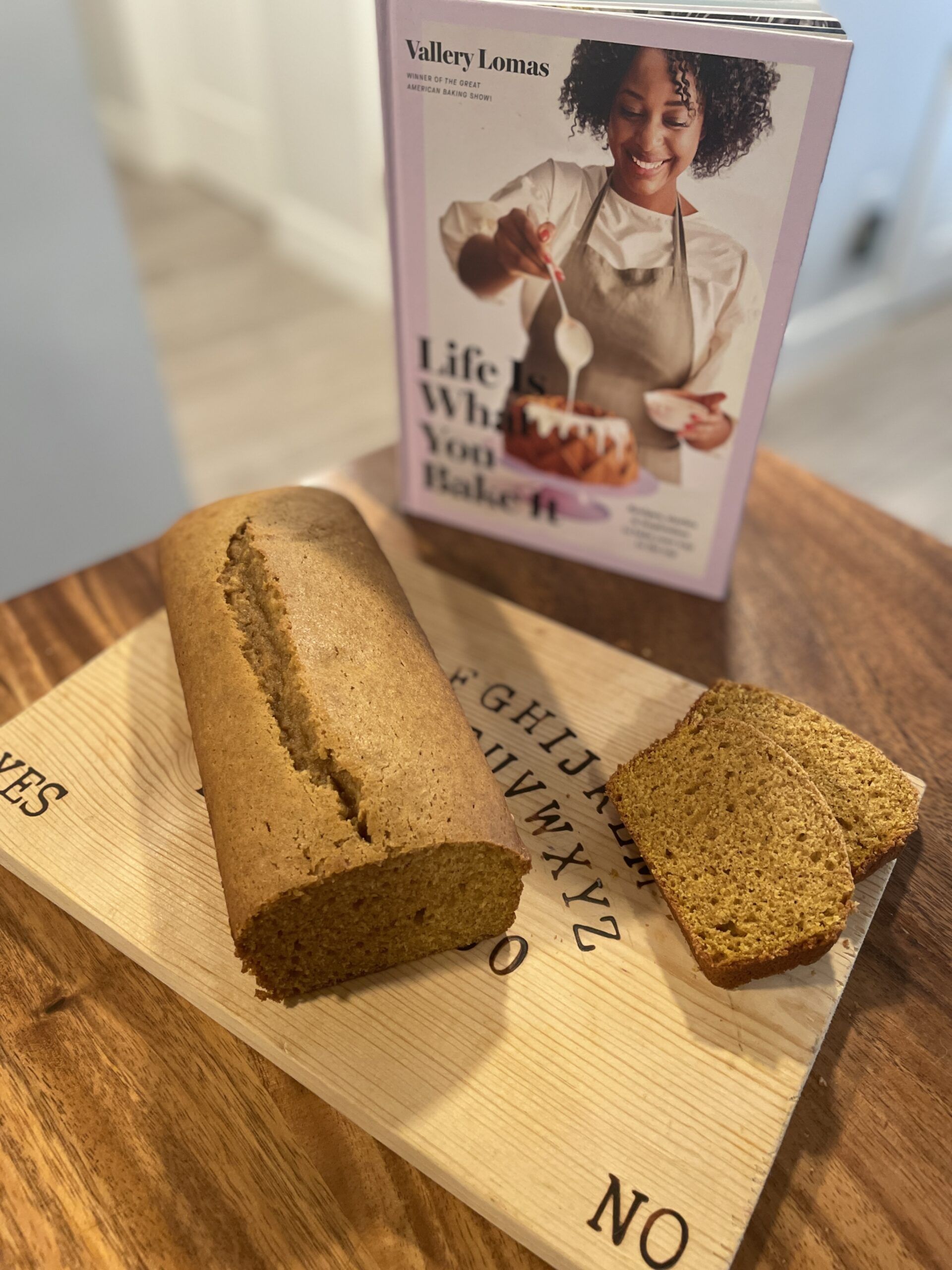 A loaf of pumpkin bread sits on a ouija board cutting board with two slices to the side on a wooden table next to the cookbook Life is What You Bake It