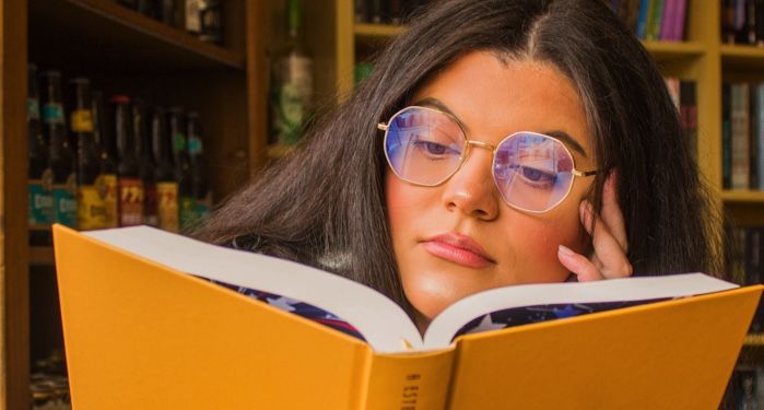 a tanned-skin woman with glasses is reading a book with a yellow cover