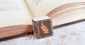 Enamel pin in the shape of a brown book with an orange leaf on it