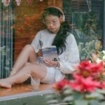 fair-skinned Asian woman reading near storefront window and flowers with her shoes off, relaxing