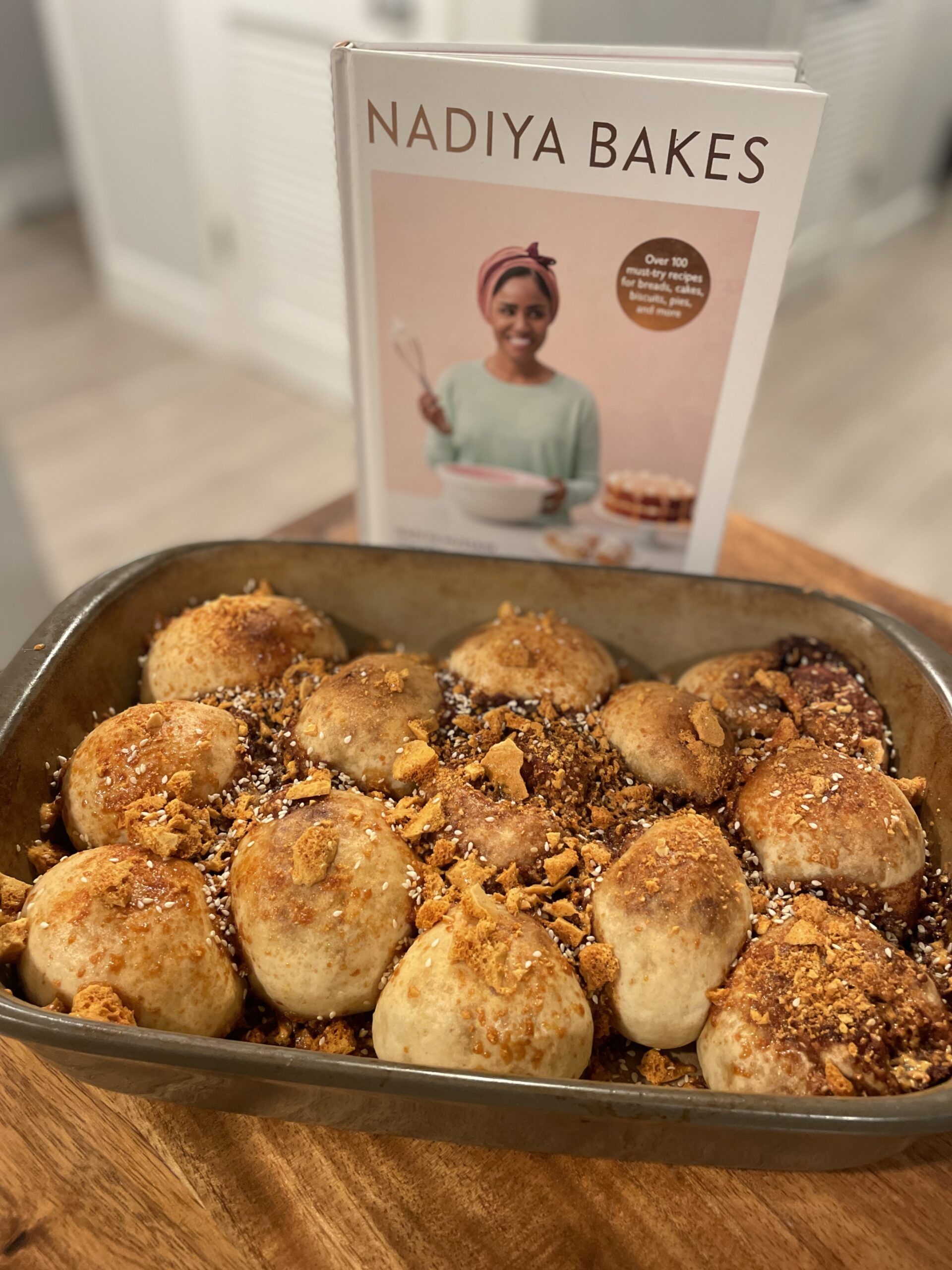 A very messy pan of mishapen rolls topped with a crumbly sugar and sesame seeds in front of the cookbook Nadiya Bakes