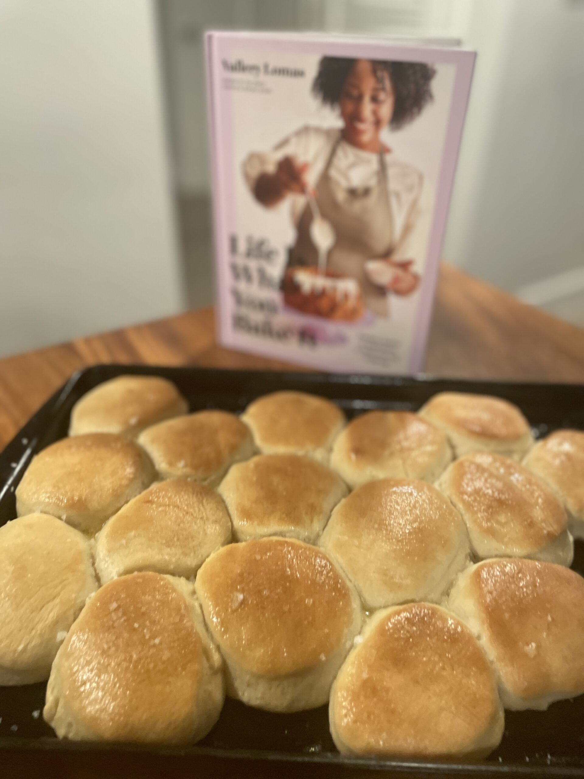 A pan of flattish dinner rolls, lightly golden, in front of the cookbook Life is What You Bake It