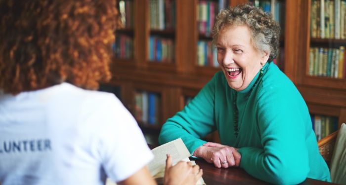 a lightly tanned skinned person with thick, curly hair is reading to a fair-skinned older woman in a library