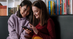 a photo of two young women in front of a bookcase looking at a phone together