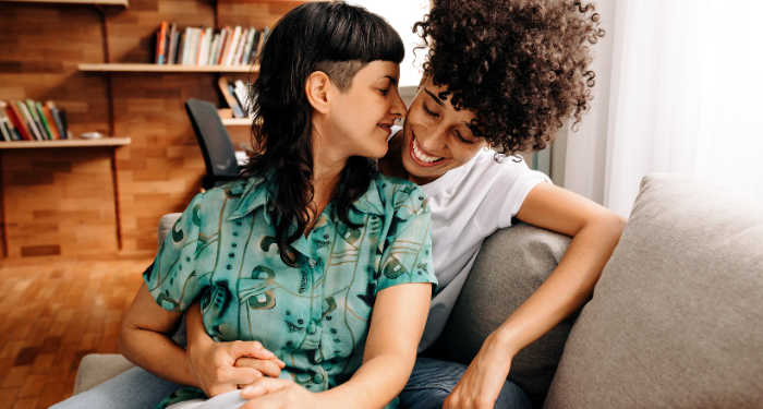 a photo of a queer couple cuddling and smiling in front of a bookcase