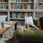 a photo of a woman reading on a couch with bookshelves in the background and stacks on books on a table