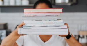 tan-skinned person is holding a stack of books in front of them, obscuring their face