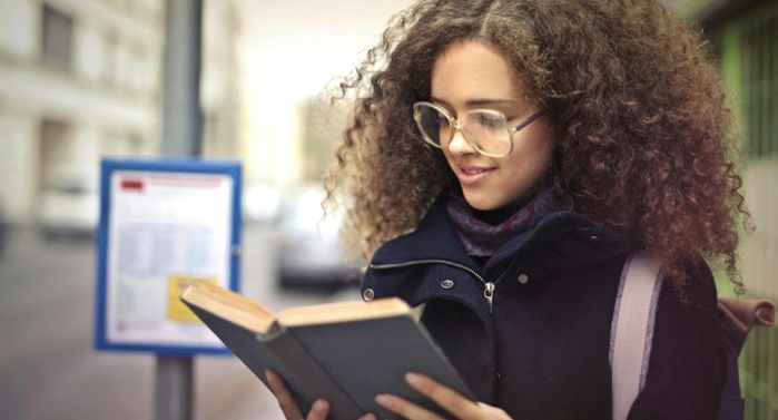 lightly-tan skinned girl with curly kinky hair reading a book at a bus stop