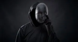 a figure dressed in a black hooded cloak with a mask in hand where one eye is visible from the holes in the mask