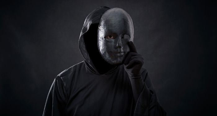 a figure dressed in a black hooded cloak with a mask in hand where one eye is visible from the holes in the mask