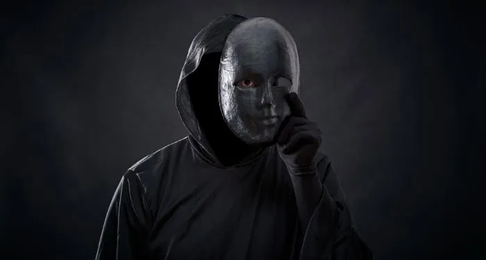 a figure dressed in a black hooded cloak with a mask in hand where one eye is visible from the holes in the mask