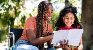 a Black woman in a wheelchair reading a book to a Black girl outside