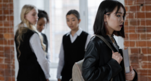 four high school students. In the foreground, one is wearing a leather jacket and carrying a book. The other students are looking at them with judgement.