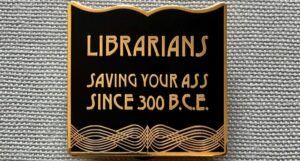 a black and gold enamel pin that says "librarians, saving your asses since 300BC"