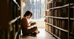 fair-skinned person reading on the floor in between library shelves