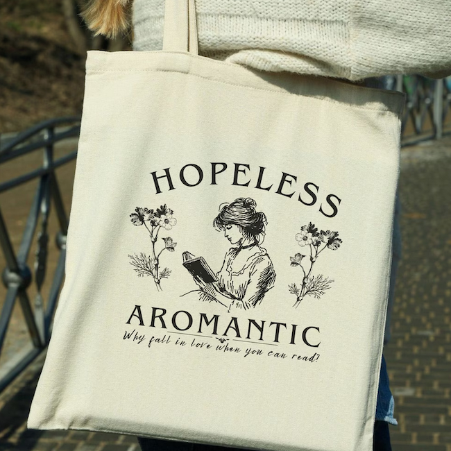 a tote bag with a black and white sketch illustration of a woman reading with text saying hopeless aromantic why fall in love when you can read?