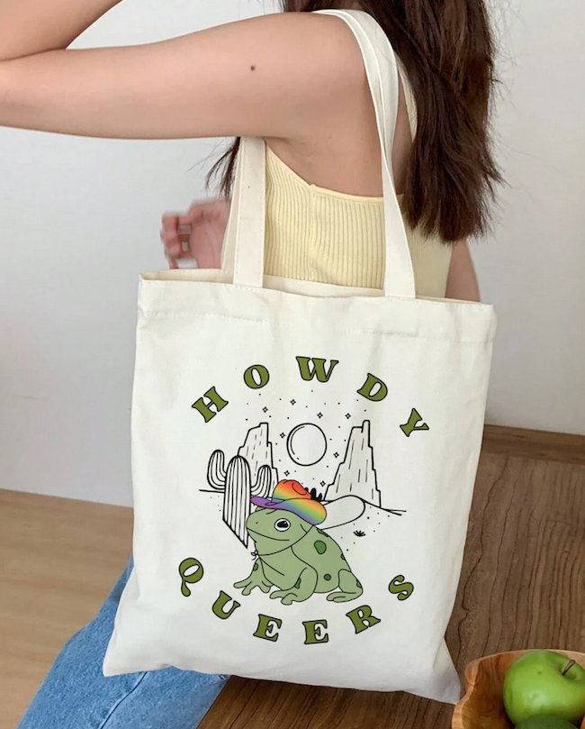 a tote bag with a graphic illustration of a frog in a rainbow cowboy hat that says howdy queers